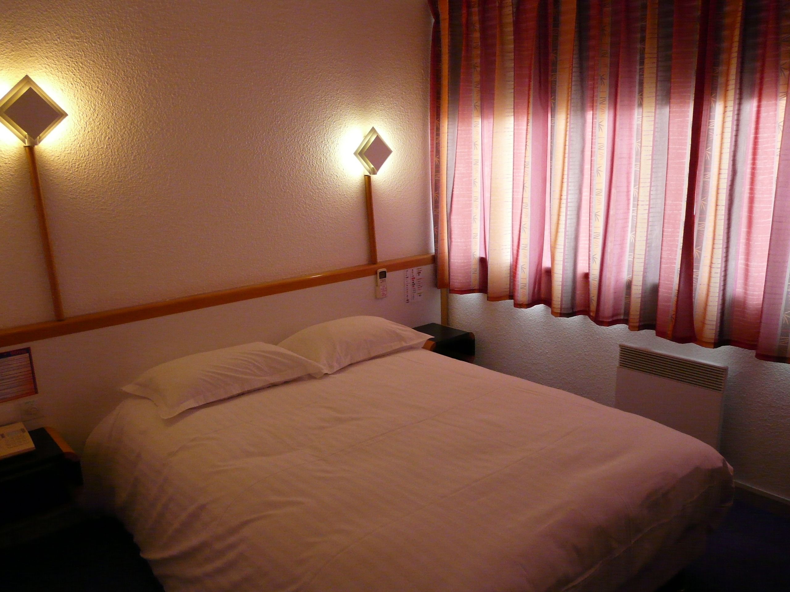 Een hotelkamer met een bed, twee wandlampen, een nachtkastje, een telefoon en een raam met gordijnen.