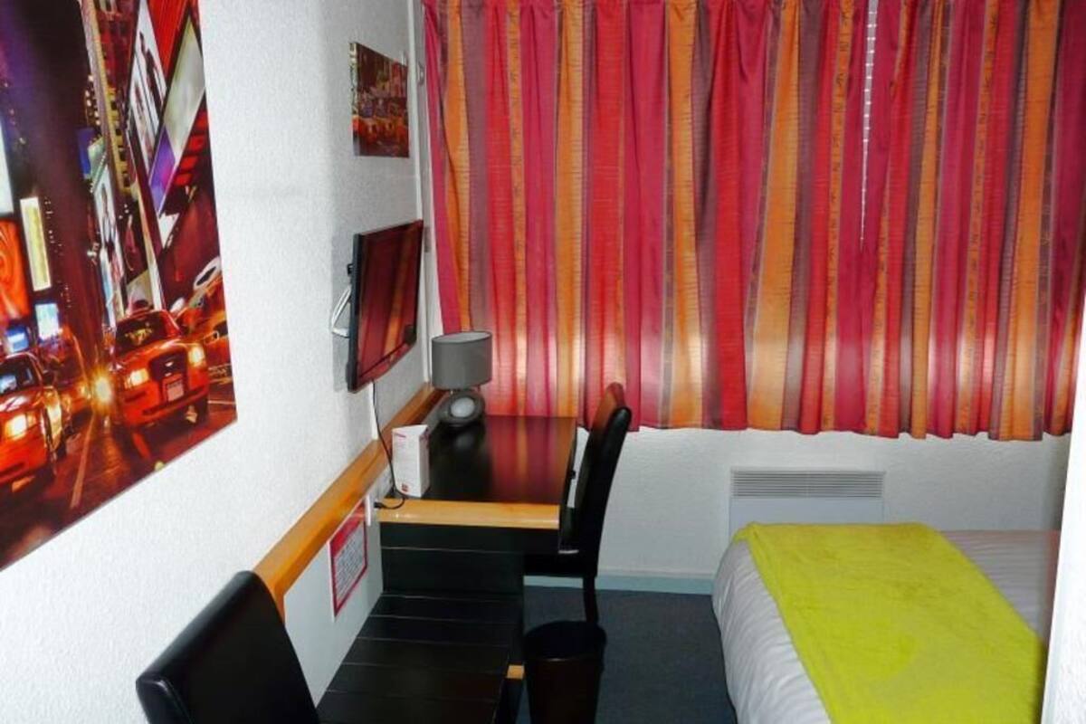 A hotel room with a bed, a desk with a chair, a lamp, a picture on the wall, and a window with curtains.