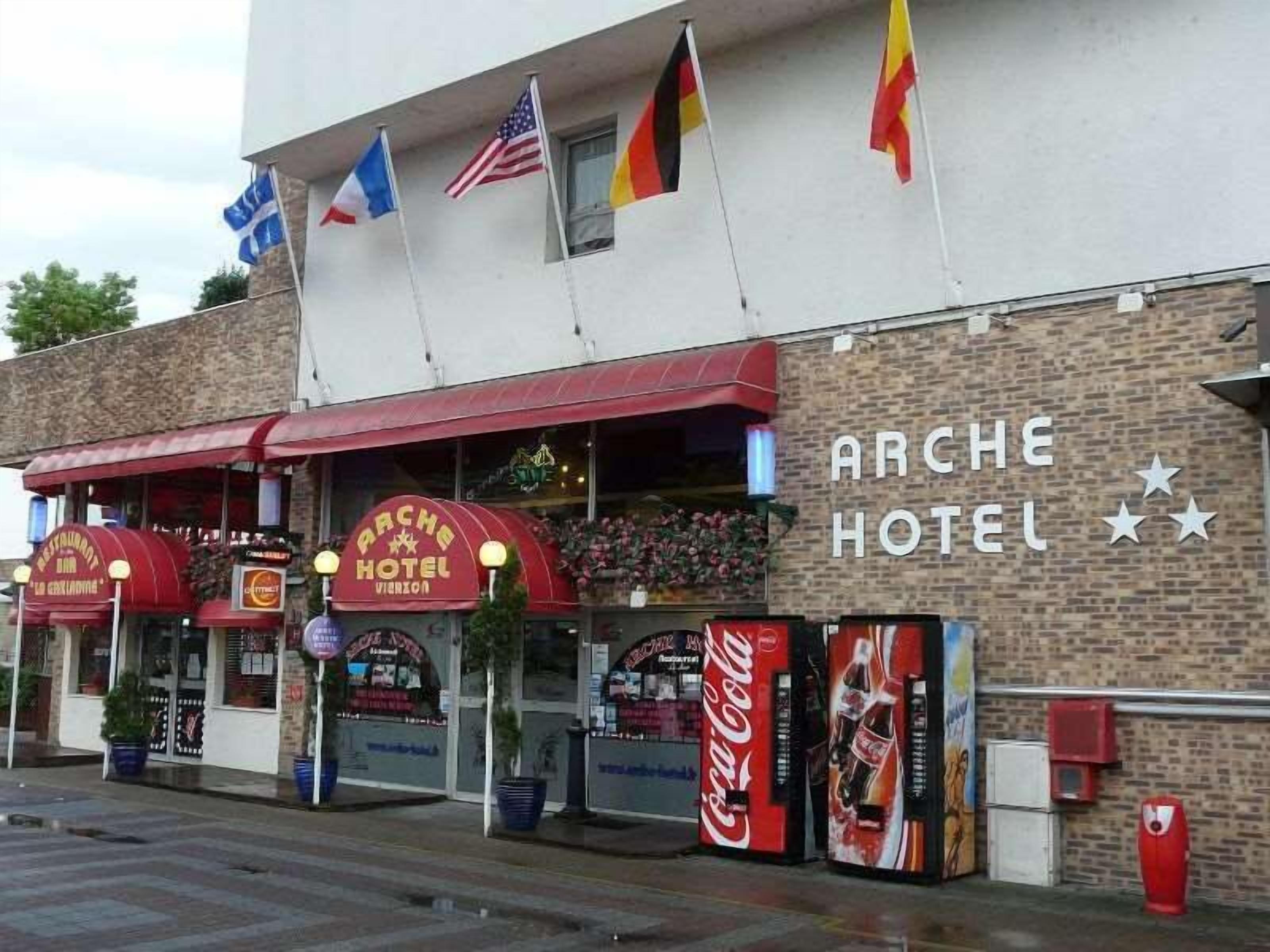 The exterior of the Arche Hotel with a red awning, multiple flags, and Coca-Cola vending machines.