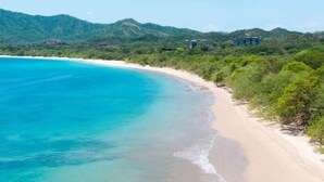 A beach with clear turquoise waters, sandy shore, and lush green hills in the background. - Seis Playas Hotel (Tamarindo)