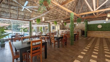Un comedor amplio con mesas y sillas de madera, techo de paja y plantas colgantes.
