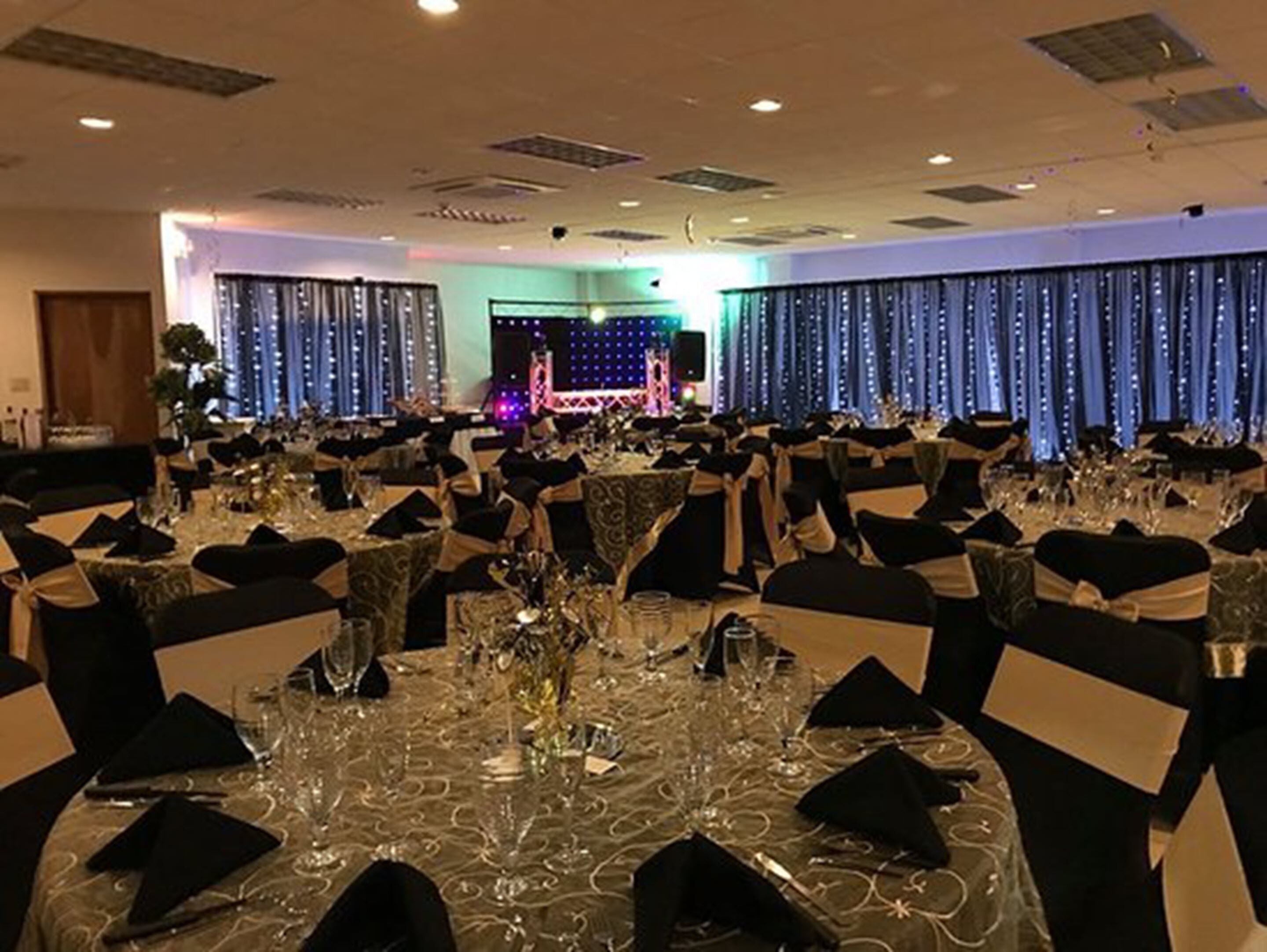 A banquet hall with round tables set for a formal event, featuring black chairs with white sashes, clear glassware, and a stage with a blue backdrop in the background.
