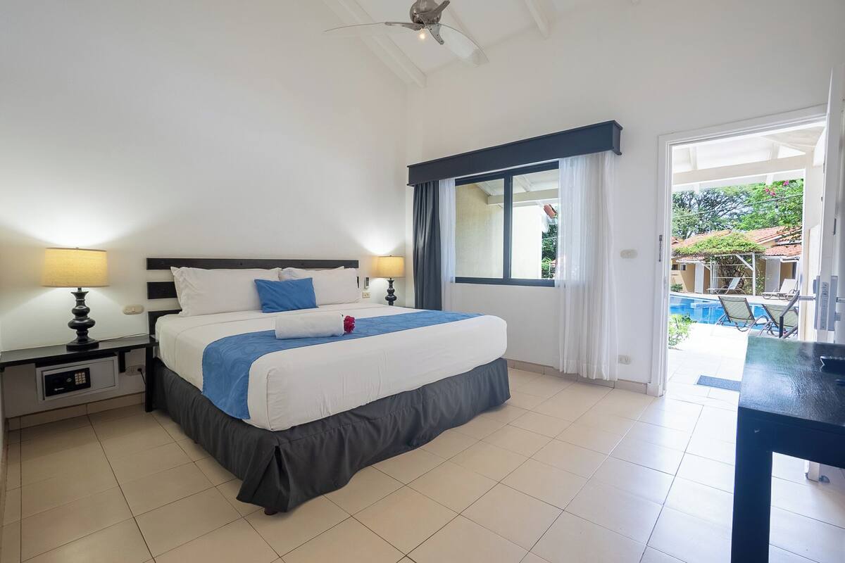 A hotel room with a bed, two bedside tables with lamps, a view of a swimming pool, and a ceiling fan.