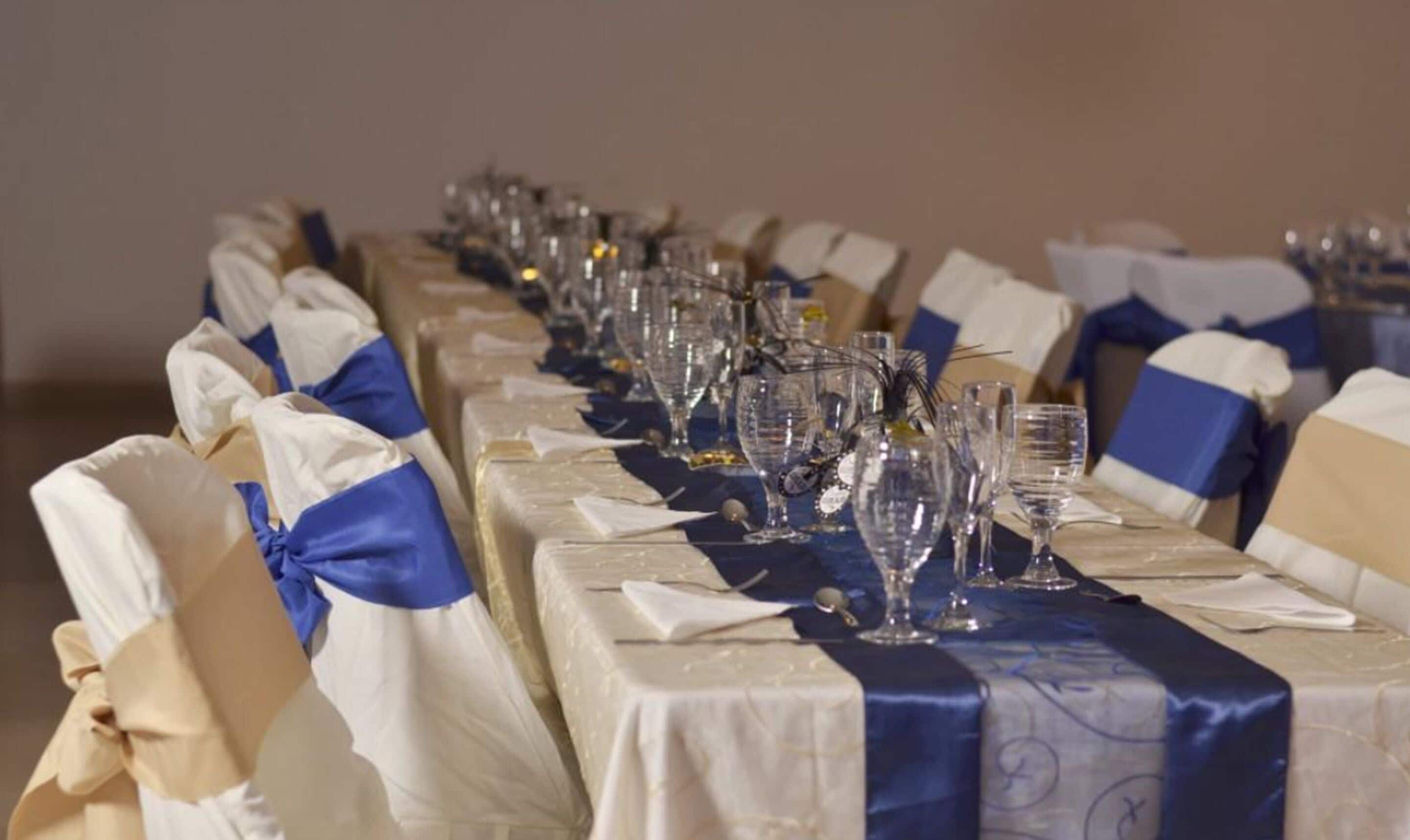 Une longue table de banquet, avec des chaises recouvertes de rubans blancs et bleus, agrémentée de verres transparents et de serviettes.