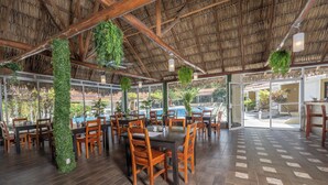An indoor dining area with wooden tables and chairs, a thatched roof, and hanging plants. - Seis Playas Hotel (Tamarindo)