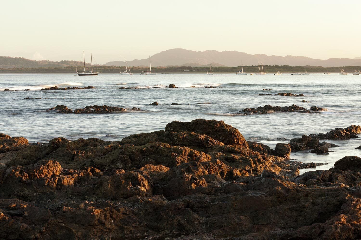 A rocky shoreline with boats in the distance and mountains in the background.