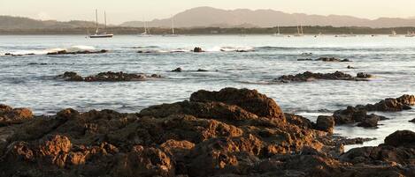 A rocky shoreline with boats in the distance and mountains in the background.