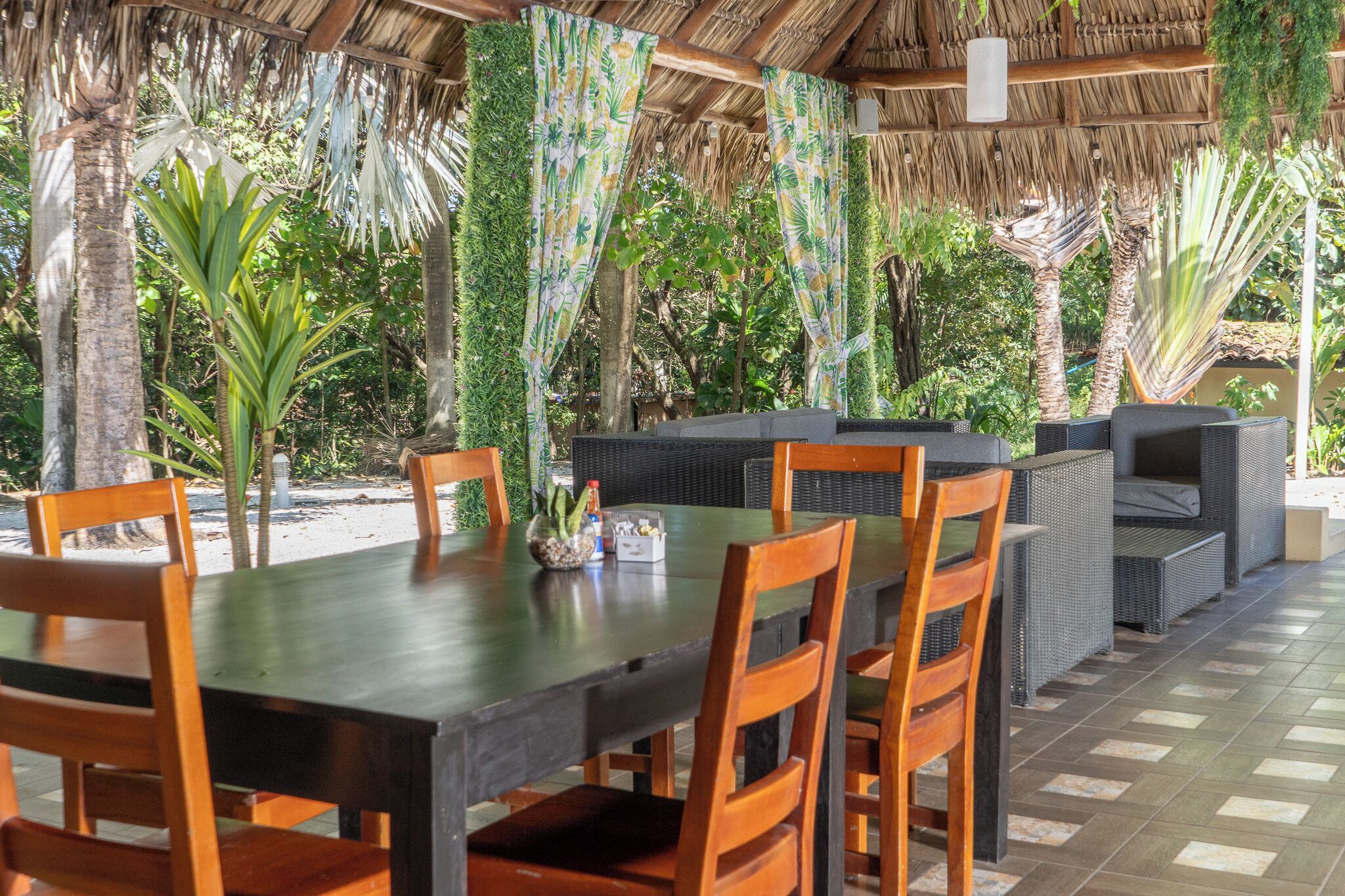 Un espace extérieur abrité, aménagé pour les repas, avec une table et des chaises, entouré d’une végétation luxuriante et surmonté d’un toit de chaume.