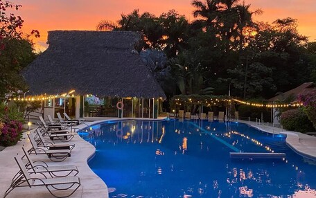 A swimming pool with lounge chairs and a thatched-roof structure, surrounded by trees and string lights.