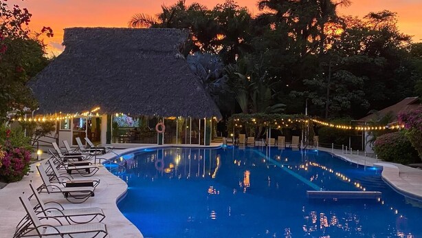 A swimming pool with lounge chairs and a thatched-roof structure, surrounded by trees and string lights. - Seis Playas Hotel (Tamarindo)