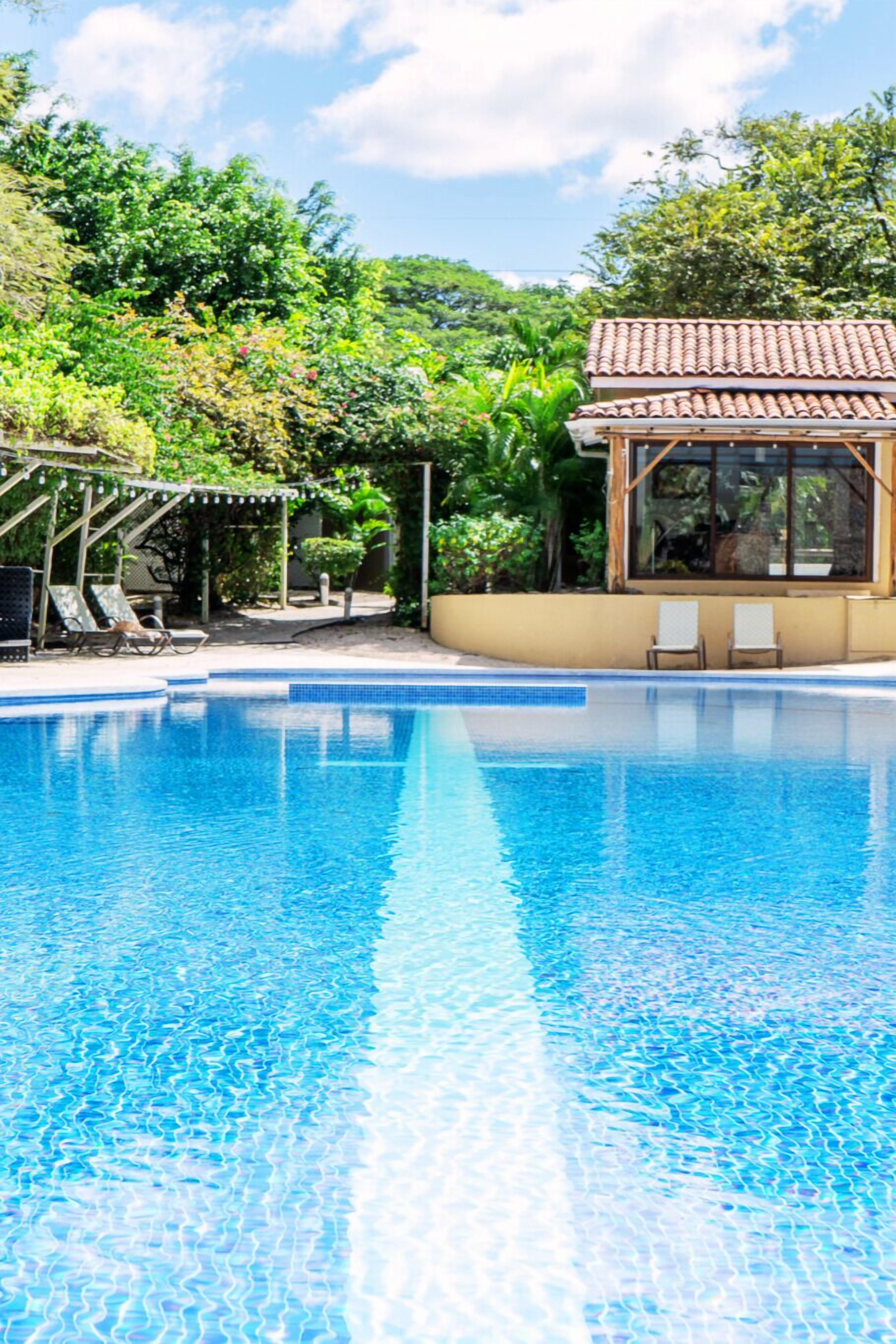 A clear blue pool with lounge chairs and a covered area, surrounded by lush greenery.