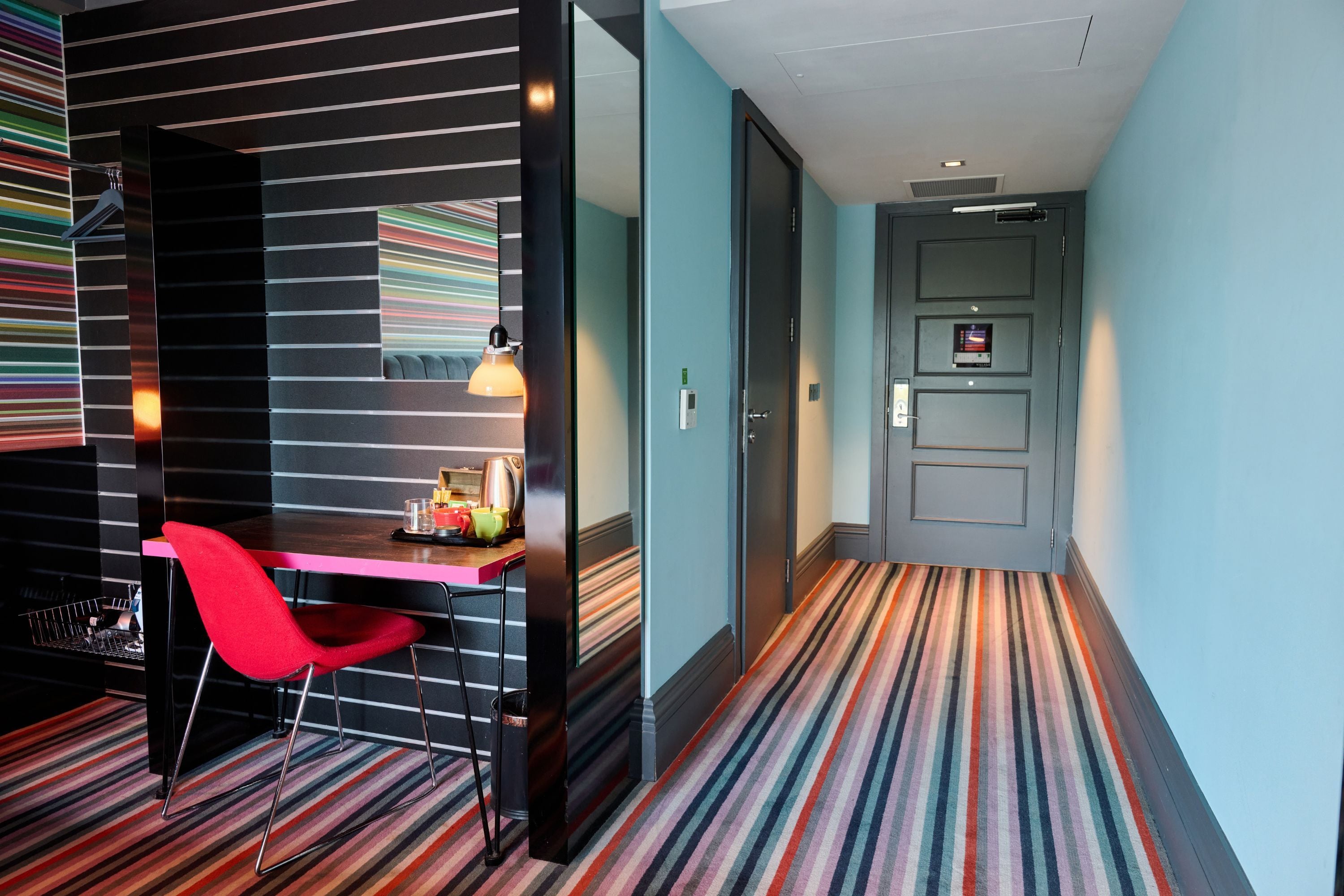 Habitación de hotel con alfombra a rayas, una silla roja, un escritorio con lámpara y un rincón americano.