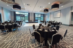 A conference room with multiple tables and chairs, large windows, and a stage. - Village Hotel London Watford (Borehamwood)