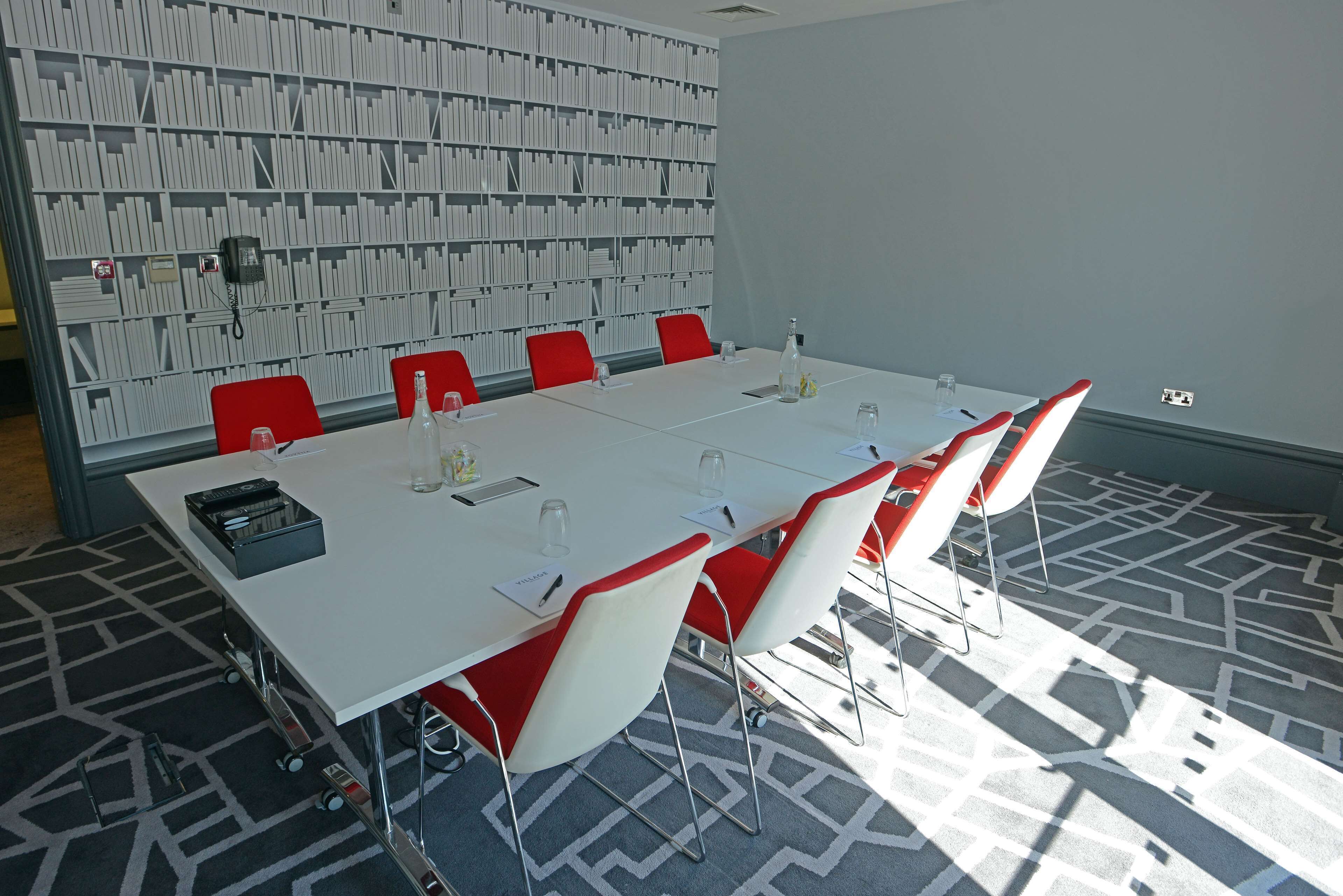 Uma sala de conferências com uma mesa branca, cadeiras vermelhas, uma estante de livros ao fundo e um tapete estampado.