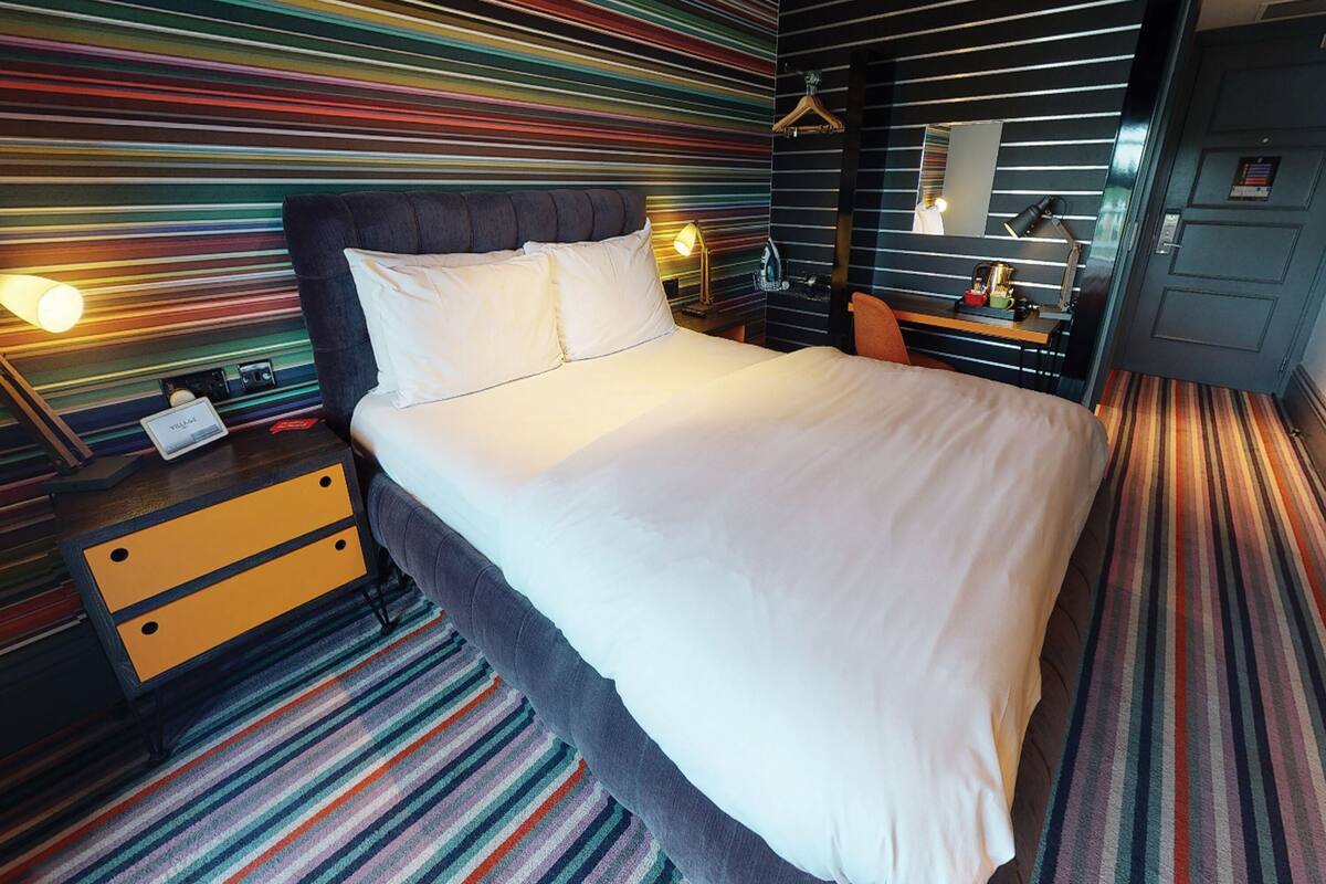 Une chambre d’hôtel avec un grand lit, une table de chevet avec une lampe, une commode et un tapis à rayures.