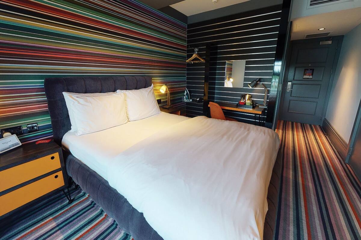 Une chambre d’hôtel avec un grand lit, une table de chevet avec une lampe, une commode et un tapis à rayures.