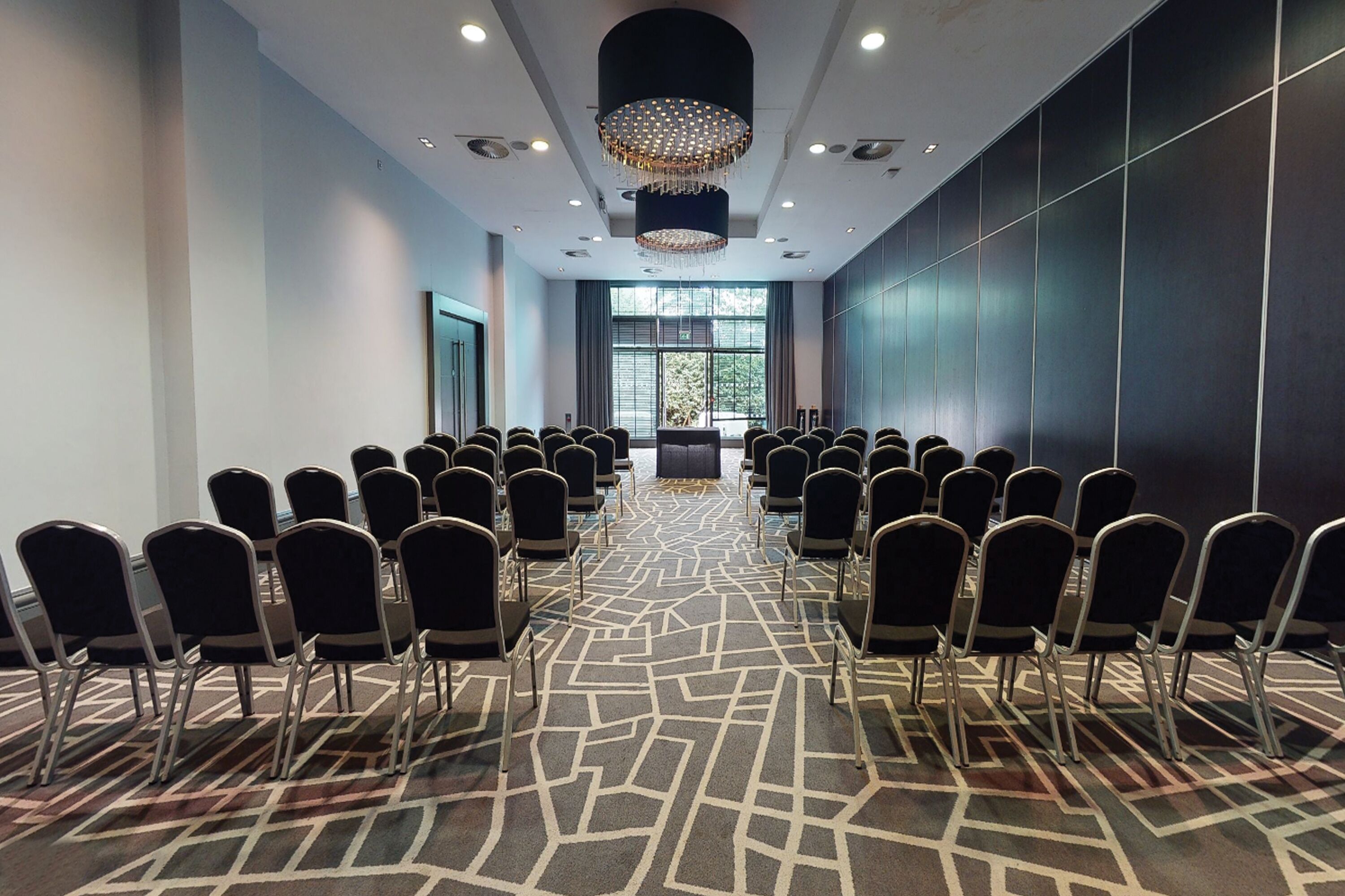 A conference room with rows of chairs facing a stage, large windows, and a patterned carpet.