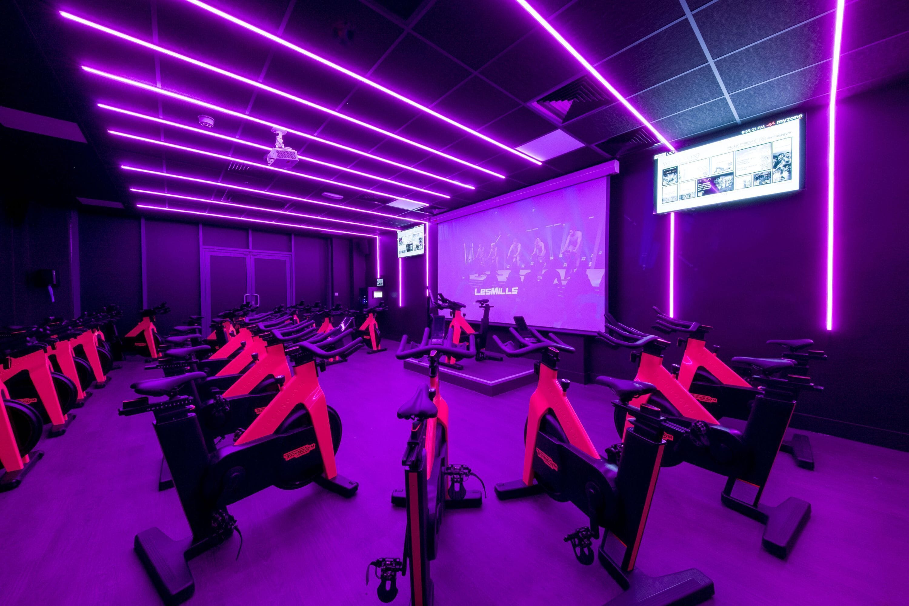 Un gimnasio con bicicletas estáticas, iluminación morada y una pantalla de proyección en la pared.