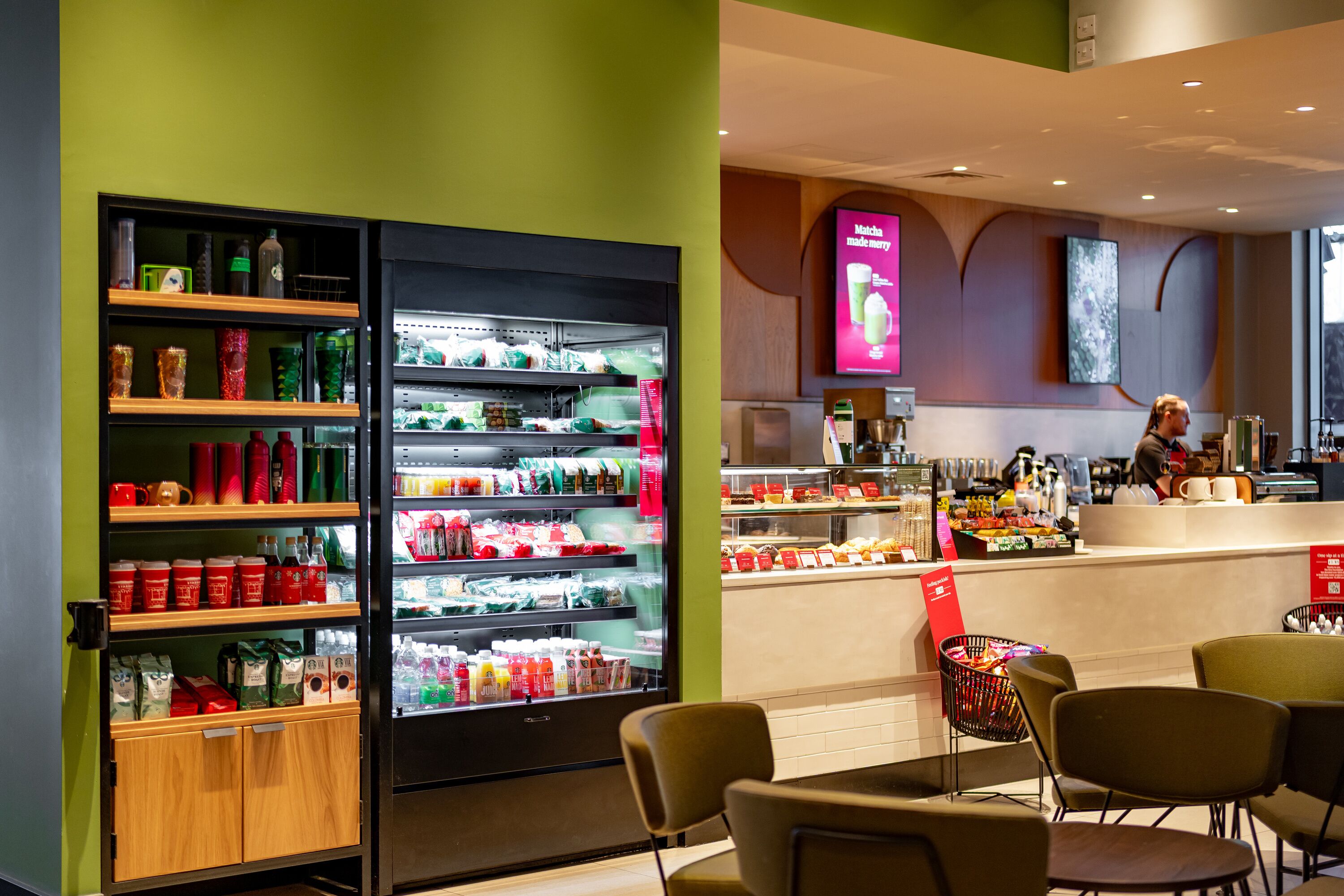Un café moderno con una paleta de colores verde y marrón, que incluye un mostrador refrigerado, una zona de estar y una barra con una persona detrás.
