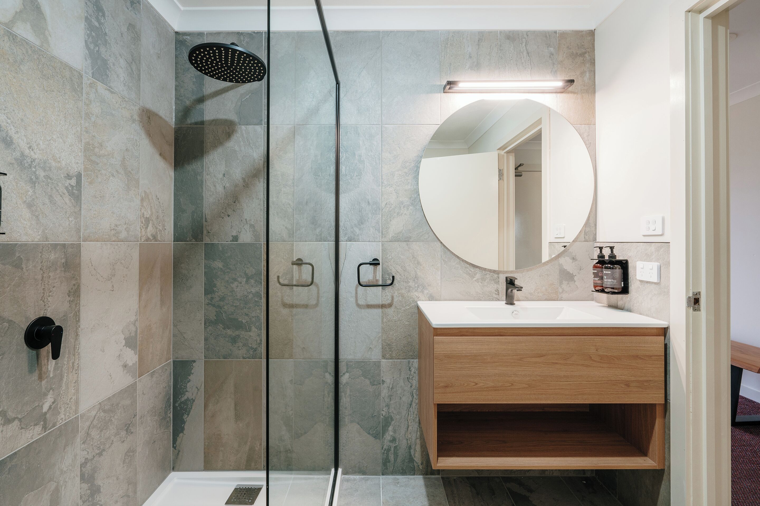 A modern bathroom with a glass-enclosed shower, a wooden vanity, and a round mirror.