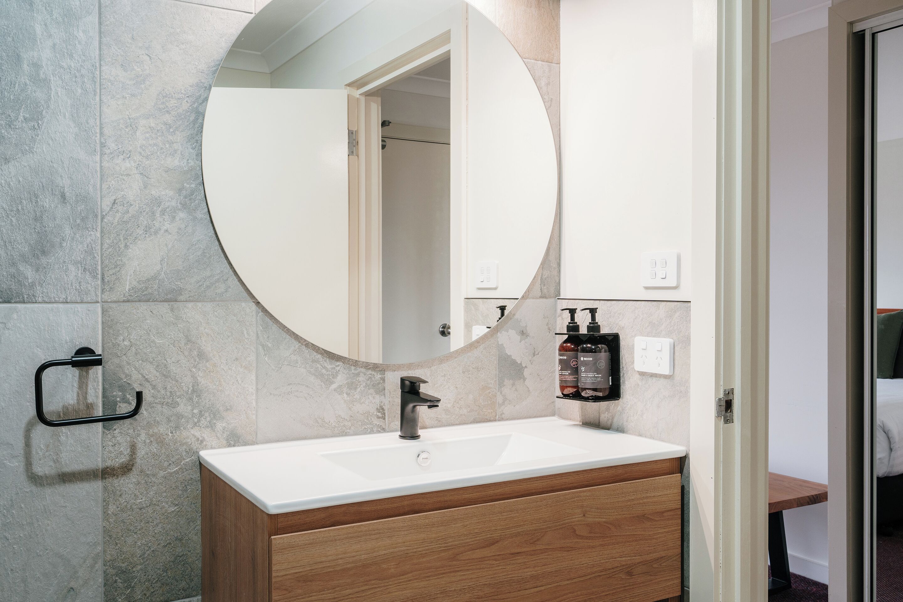 A bathroom with a round mirror, a white sink, wooden cabinetry, and a towel rack.