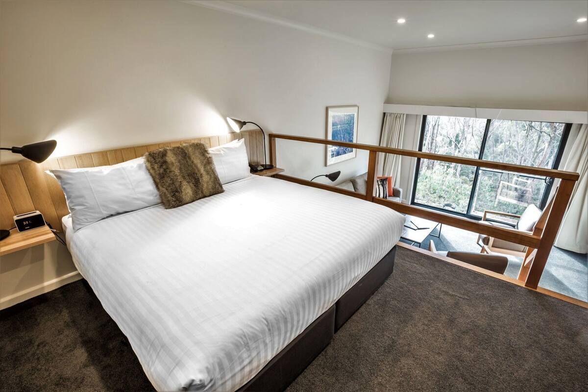 Ein modernes Hotelzimmer mit einem großen Bett, einem Holz-Kopfteil, einem Nachttisch mit Lampe sowie einem Blick ins Freie durch einen Balkon und ein Fenster mit Vorhängen.