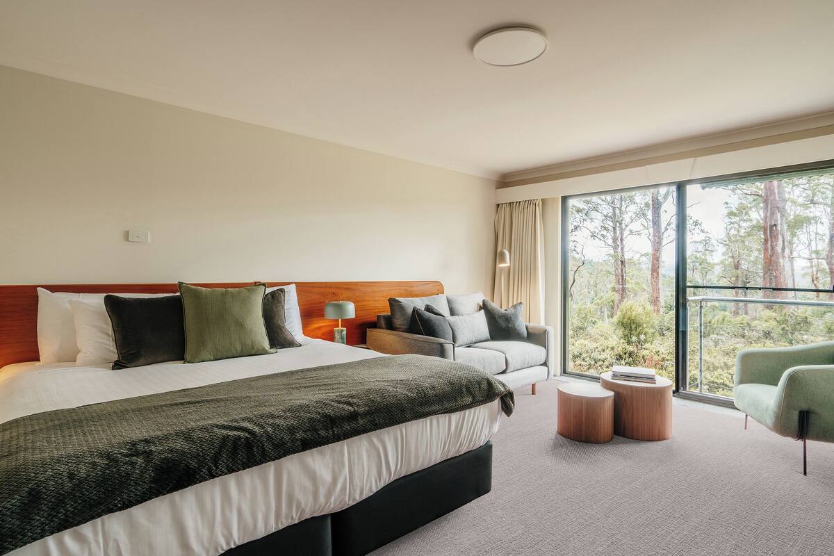 Ein modernes Hotelzimmer mit einem großen Bett, einer Sitzecke und Blick auf Bäume durch große Fenster.