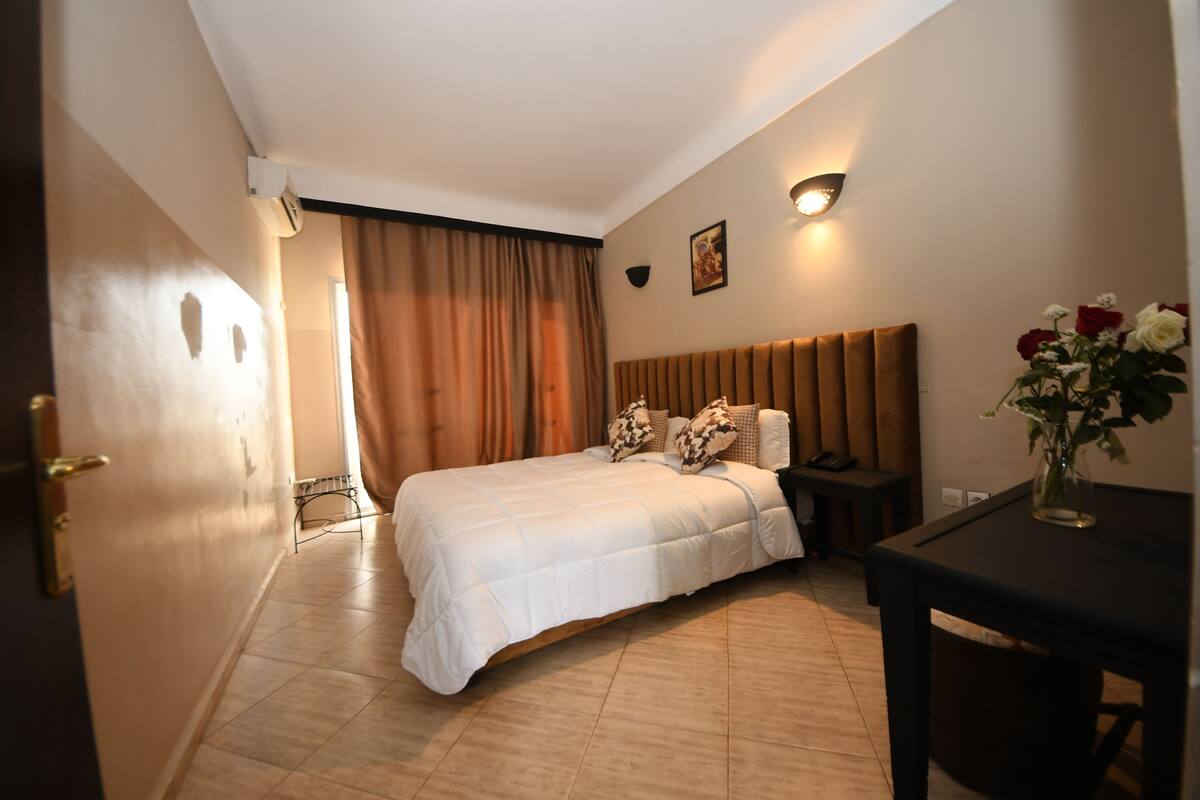 A hotel room with a bed, bedside table, and a small table with a vase of flowers.