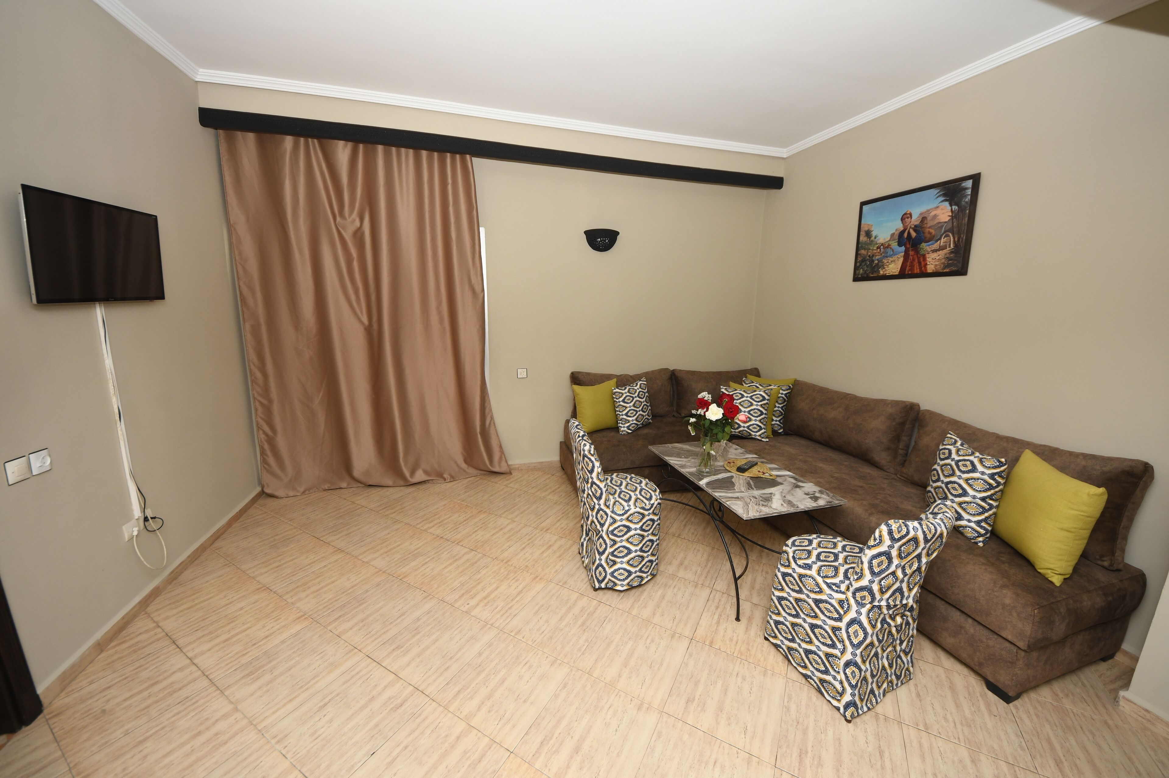 Una sala de estar con un sofá marrón, sillones estampados, una mesa de centro y un televisor montado en la pared.