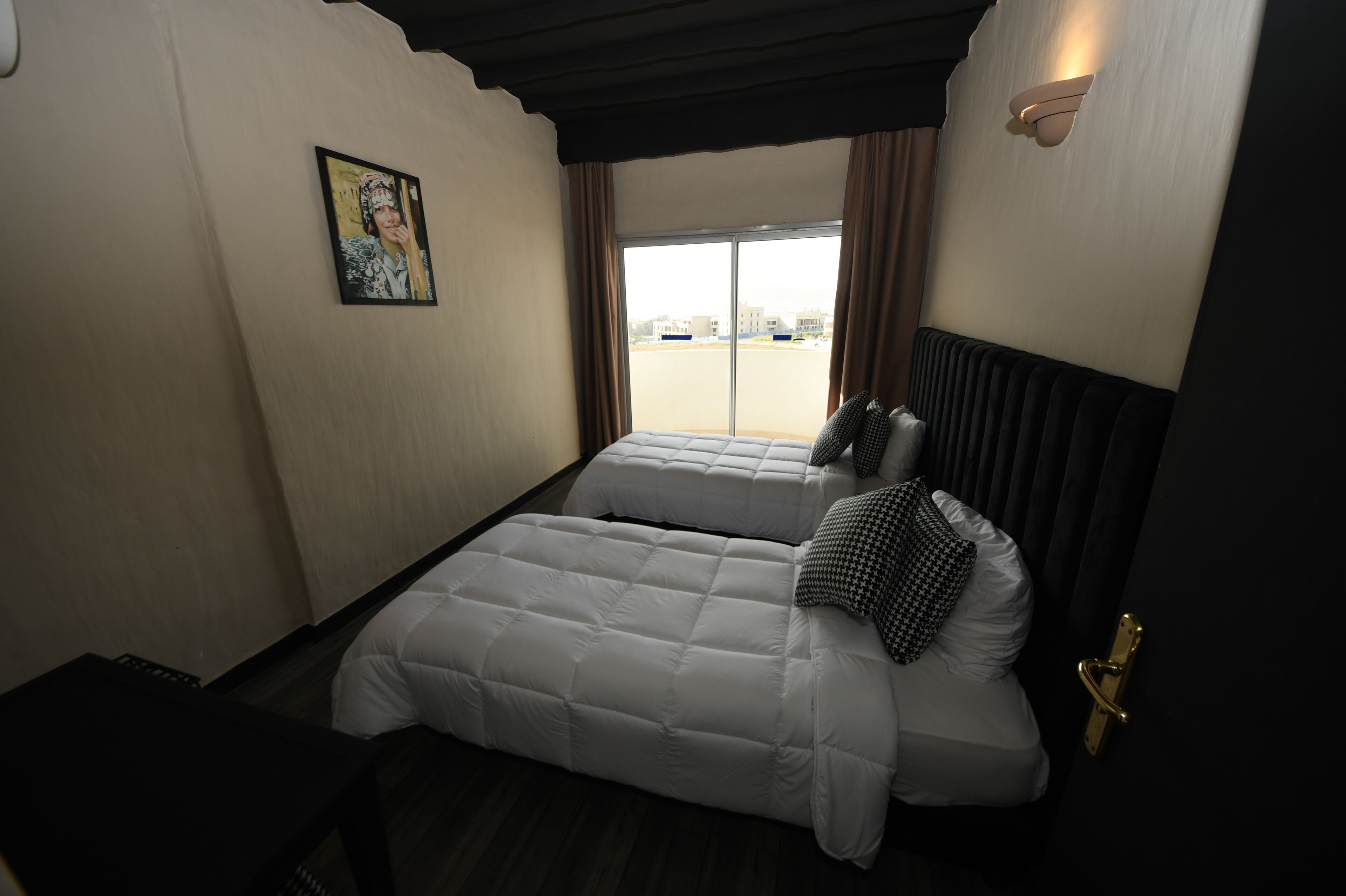 Habitación de hotel con cama, ventana con cortinas, lámpara de pared y puerta con pomo dorado.