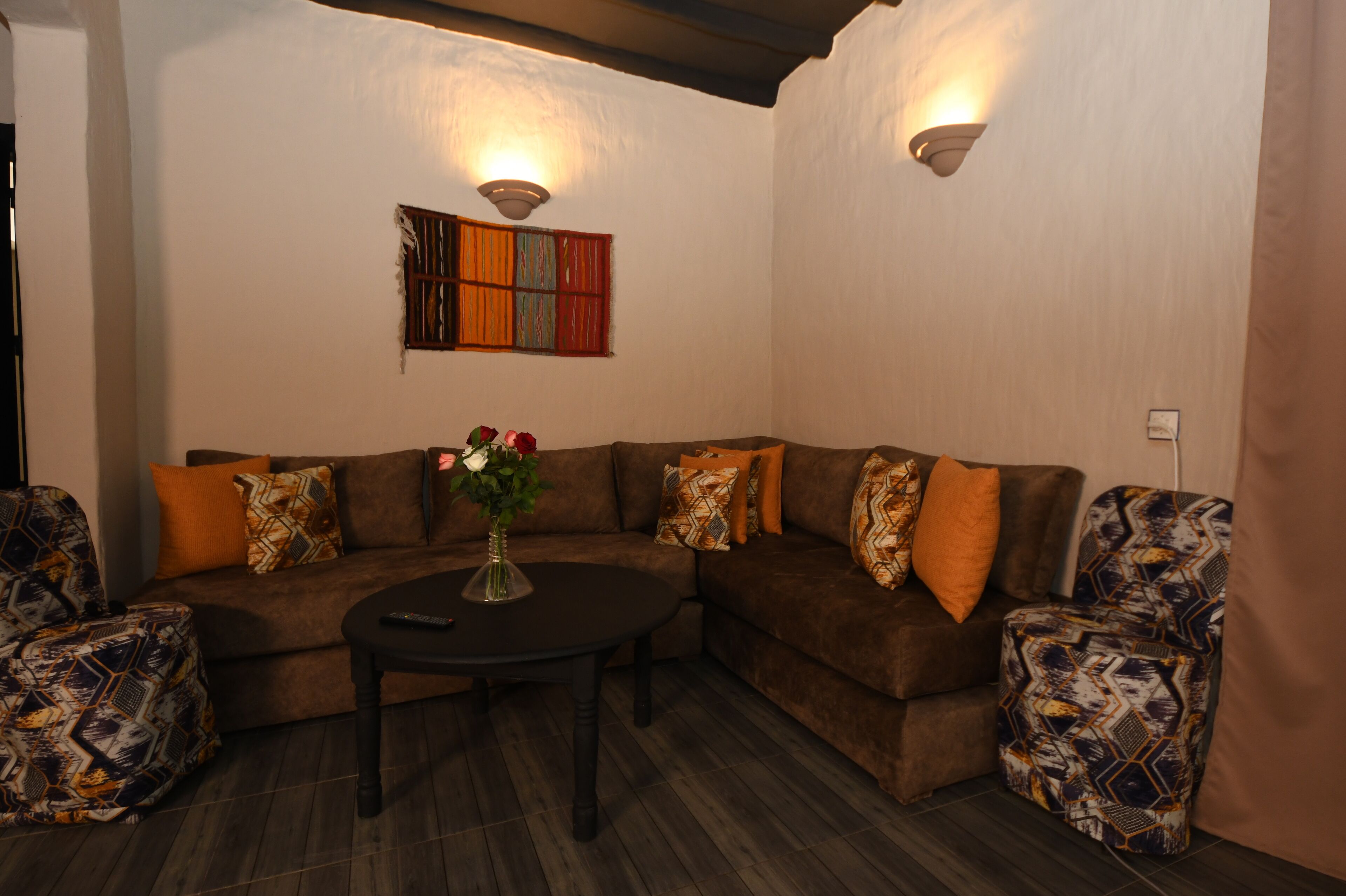 A living room with a brown sectional sofa, patterned armchairs, a round coffee table, and a vase with flowers.