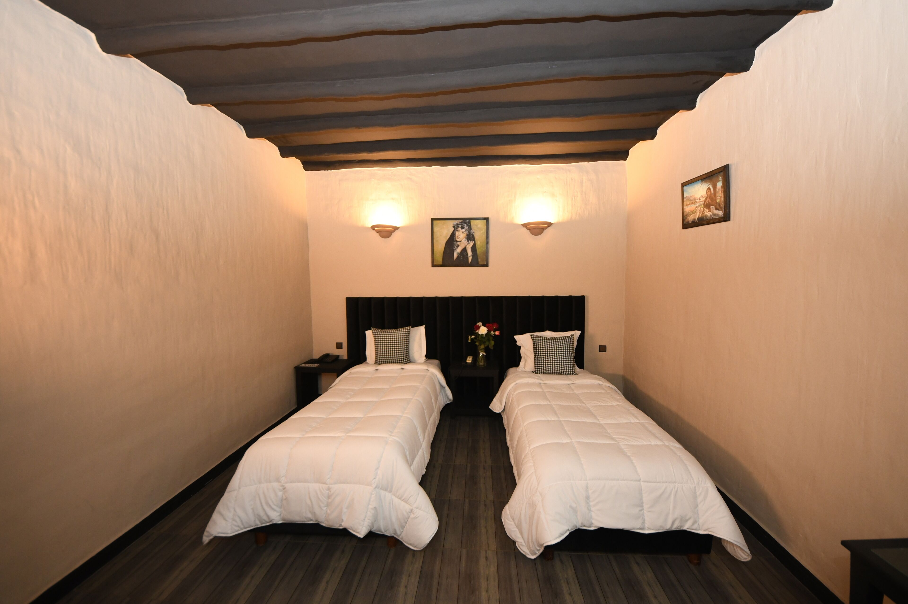 A hotel room with two beds, wooden flooring, a dark headboard, wall-mounted lights, and a framed picture.
