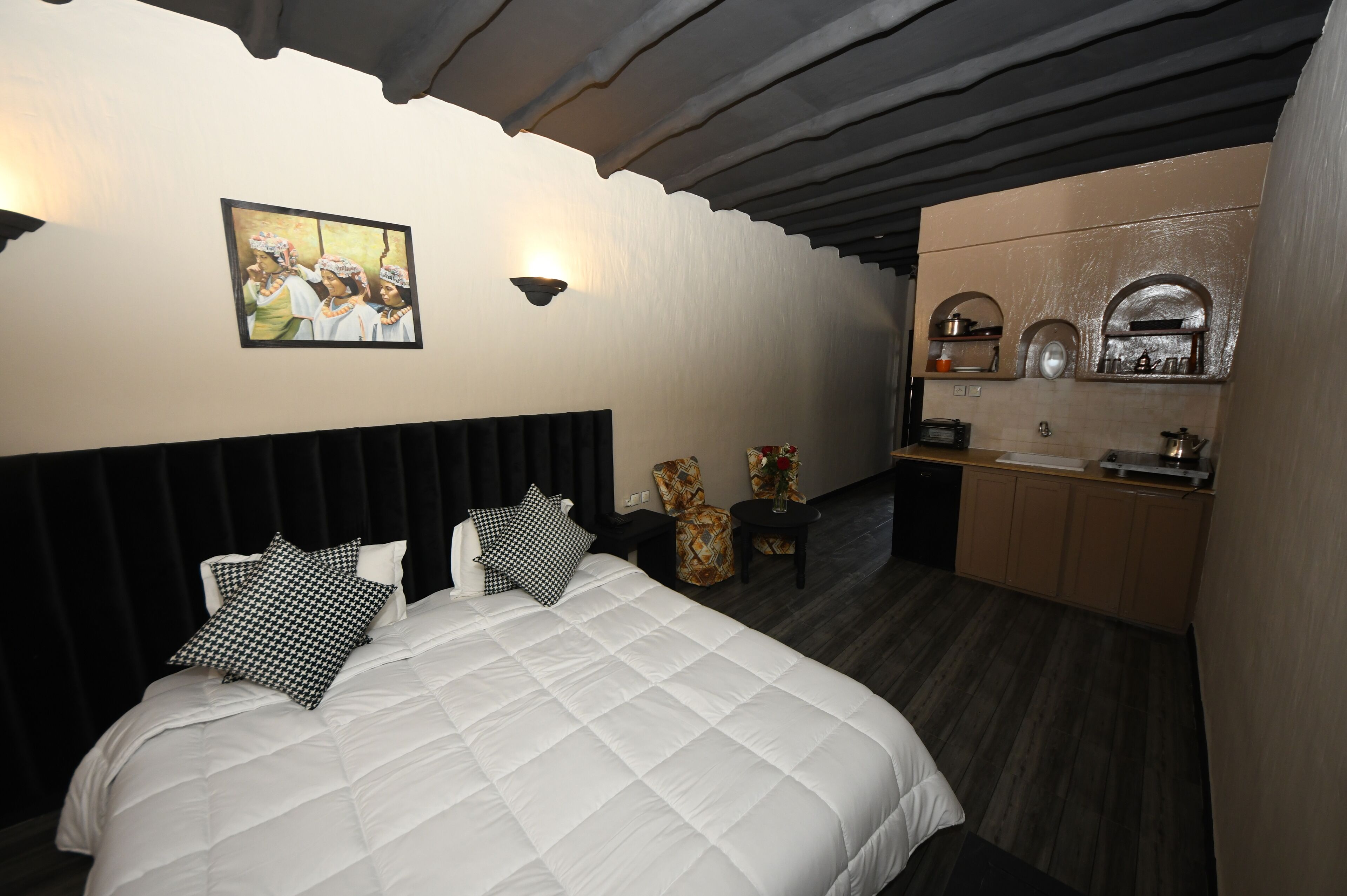 A bedroom with a large bed, a headboard, a wall-mounted picture, a small table with a vase, and a kitchenette area with cabinets and a sink.