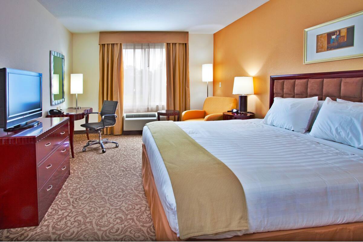 A hotel room with a large bed, a desk with a television, a chair, and a lamp.