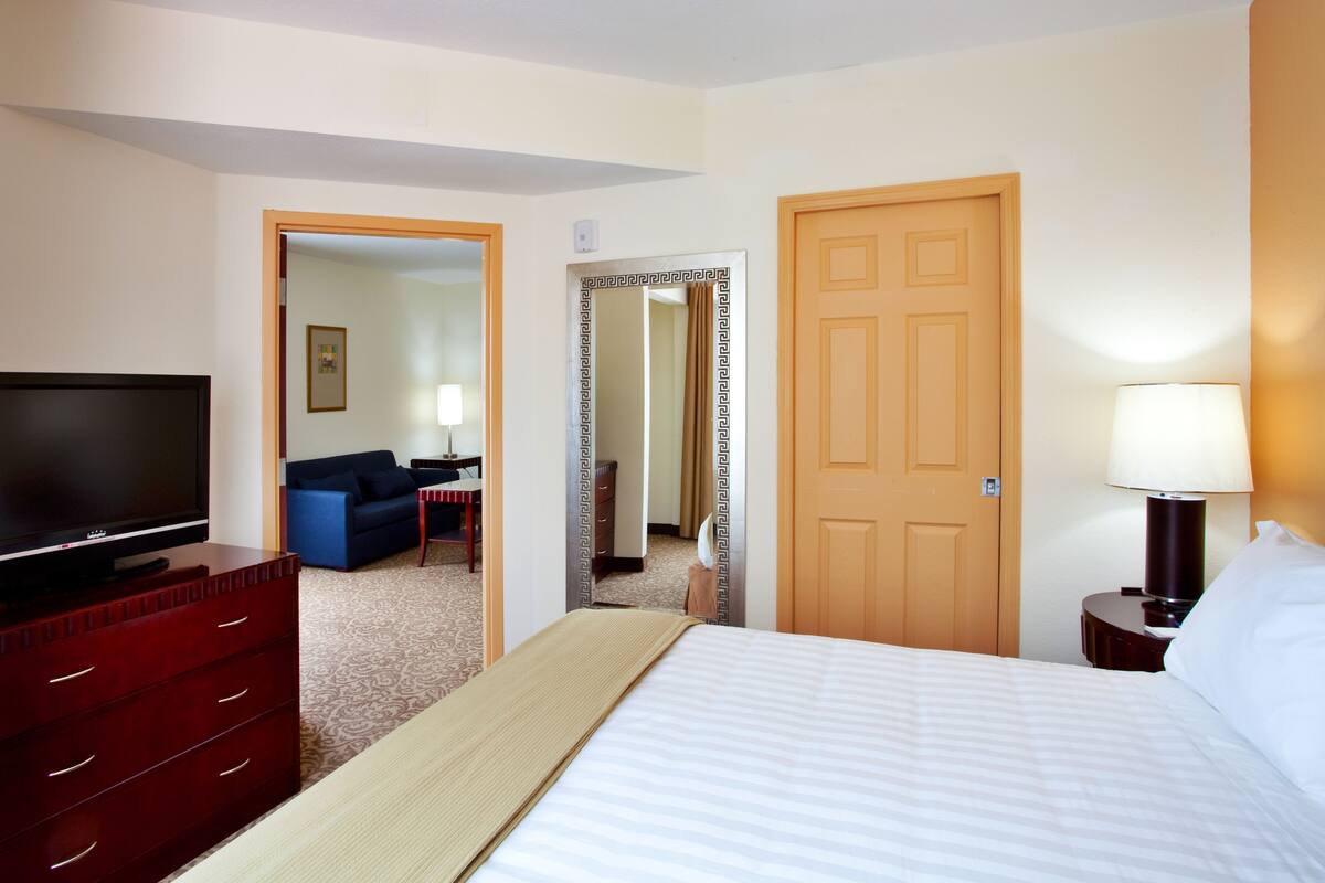 A hotel room with a bed, a TV, a dresser, a lamp, and a door leading to another room.