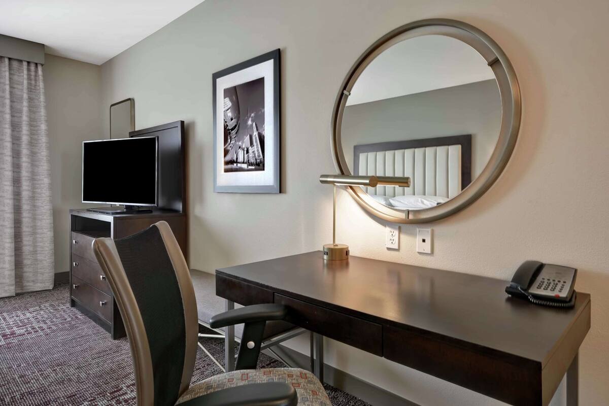A hotel room with a desk, chair, television, framed picture, round mirror, and telephone.