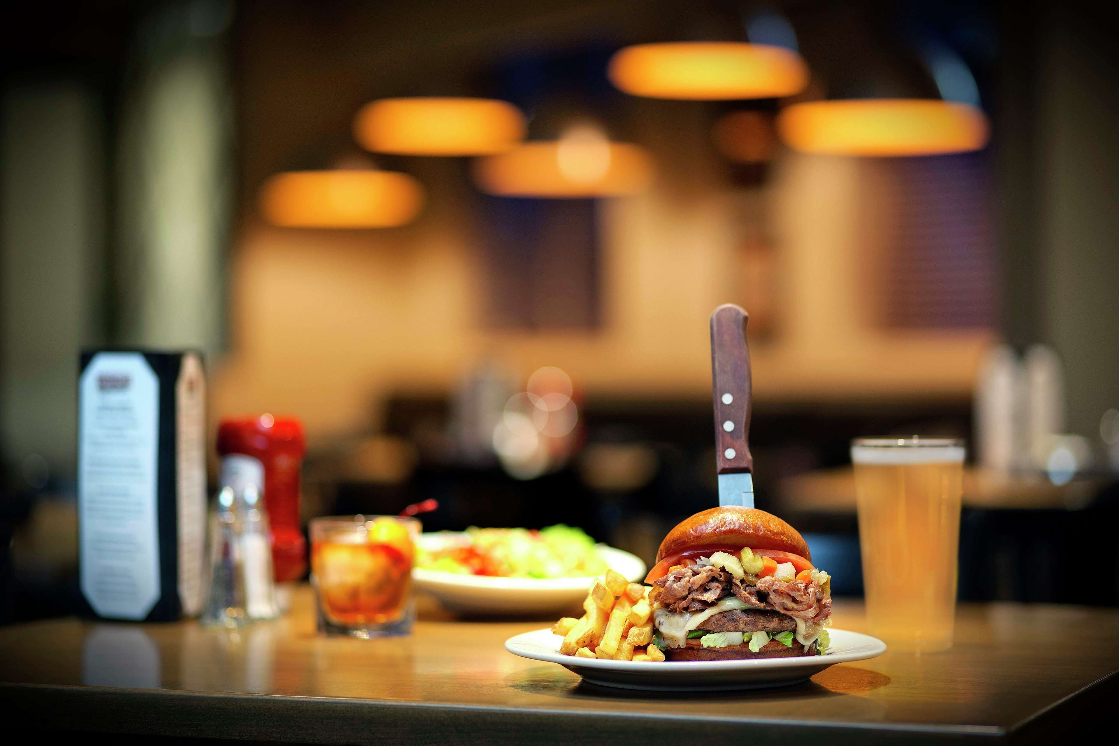 Un hamburger avec un couteau posé dessus, un verre de bière et une assiette de frites sur une table.