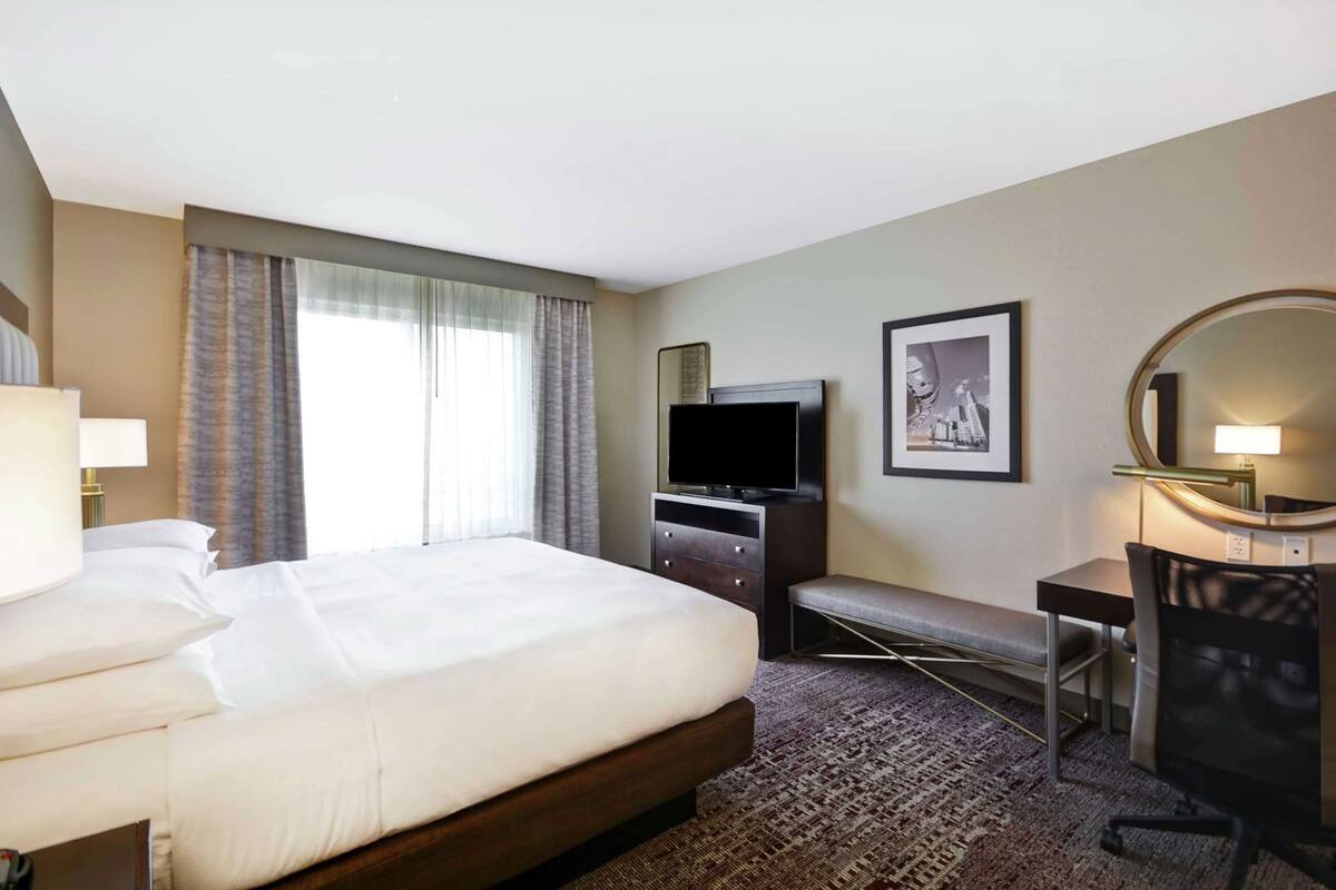 A hotel room with a large bed, a desk with a chair, a television, a desk lamp, a framed picture, and a window with curtains.