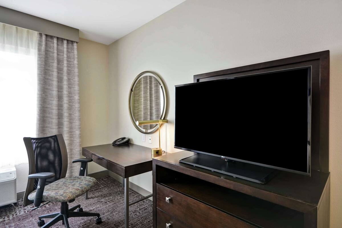 A hotel room with a desk, chair, television, and a mirror.