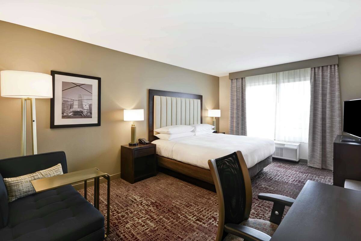 A hotel room with a bed, bedside tables with lamps, a desk, a chair, and a large window with curtains.