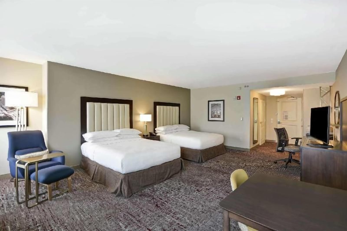 A hotel room with two beds, a desk, a chair, a television, and a carpeted floor.