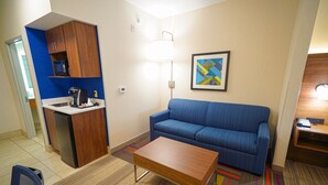 A compact hotel room with a kitchenette, a sofa, a coffee table, and a colorful rug.