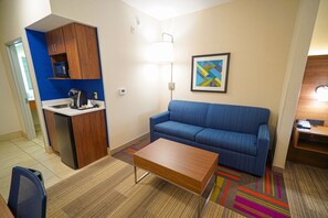 Ein kompaktes Hotelzimmer mit Küchenzeile, einer Couch, einem Couchtisch und einem farbenfrohen Teppich.