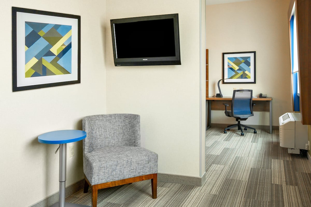 Habitación de hotel con un sillón gris, una mesita pequeña azul, un televisor de pantalla plana y dos cuadros abstractos enmarcados en la pared.