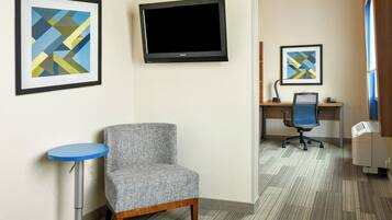 A hotel room with a flat-screen TV, a grey upholstered chair, a small blue table, and two framed abstract artworks on the wall.
