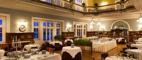 Une salle à manger spacieuse, avec de nombreuses tables dressées pour un événement formel, de grandes fenêtres et un balcon agrémenté de plantes.