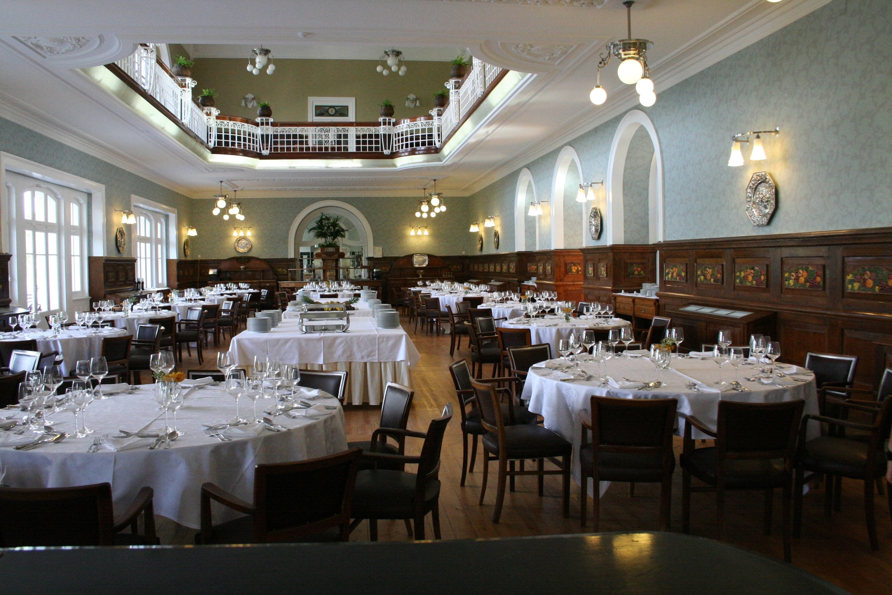 A spacious dining hall with multiple round tables set for dining, a balcony with railings, and a central decorative arch.