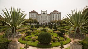 Un edificio imponente con arquitectura simétrica, un jardín formal con céspedes cuidados y setos topiarios, y una fuente central.