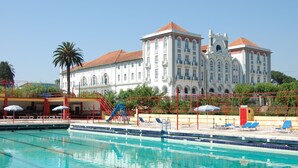 Una piscina con sillas de descanso y sombrillas, una palmera y un edificio blanco grande con techo rojo al fondo.