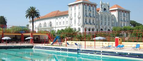 A swimming pool with lounge chairs and umbrellas, a palm tree, and a large white building with a red roof in the background.